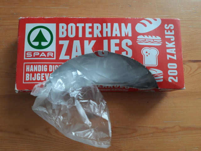 Eine rote Box mit weißer Aufschrift "Boterham Zakjes". Die Box ist geöffnet, eine durchsichtige Butterbrottüte schaut heraus.