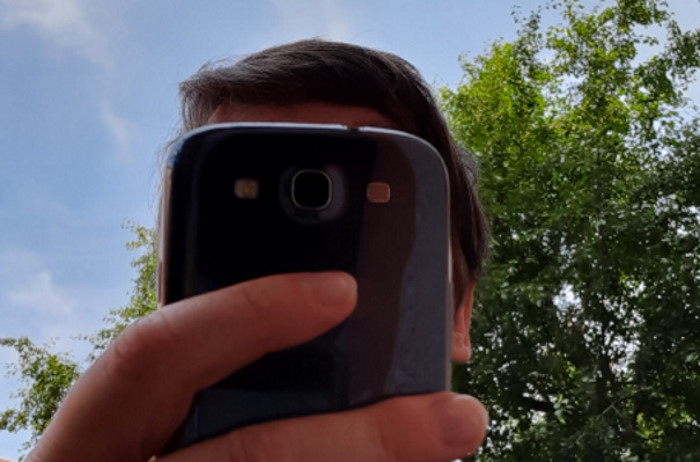Untersichtaufnahme. Eine Hand hält ein Smartphone. da Gesicht dahinter ist verdeckt. Im Hintergrund Himmel und grünes Laub.